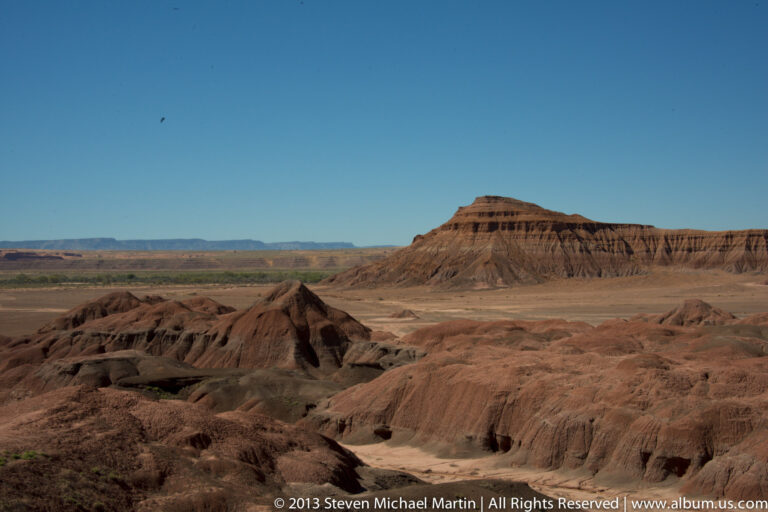 Dark eroded ridges and a pale wash fill the foreground, with a long mesa and layered plateaus beyond.