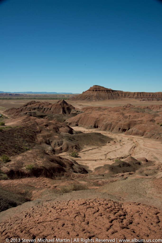 A wide desert basin of rust-colored ridges and pale washes extends toward low mesas beneath a clear sky.