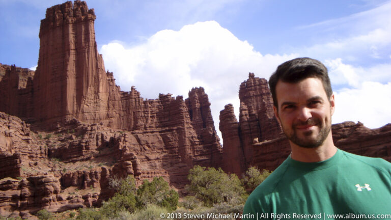 A man in a turquoise shirt poses before red sandstone fins, with desert shrubs and rocky slopes behind him.
