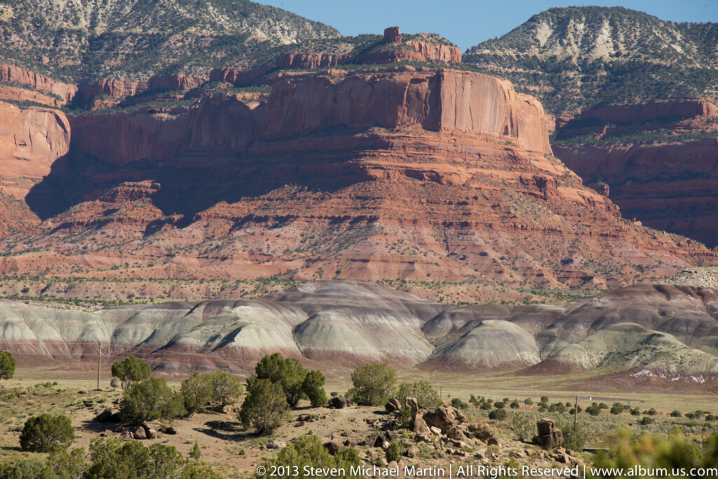 Color-banded eroded hills and scattered trees lie below a sunlit red sandstone mesa with a steep cliff face.