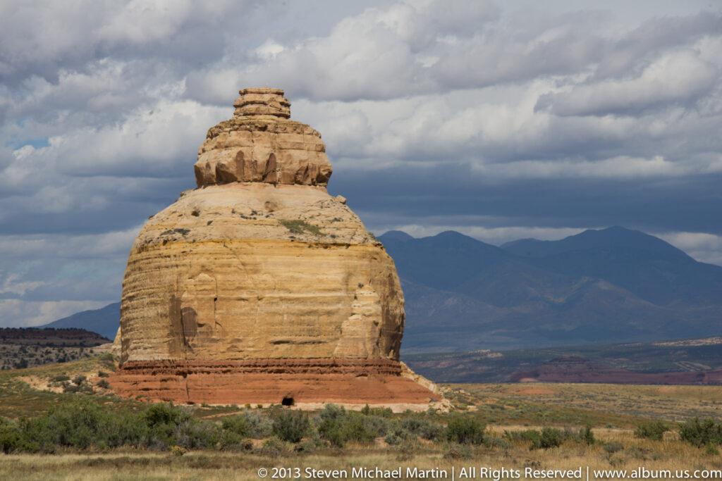 A rounded sandstone monolith with a stacked crown stands before distant blue mountains beneath thick clouds.