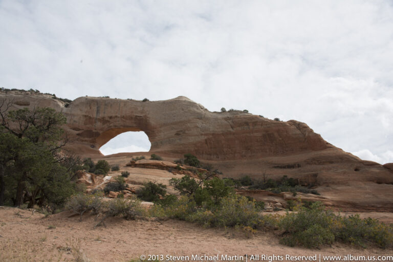 A broad sandstone arch spans a bright opening above a slope dotted with shrubs and small trees.