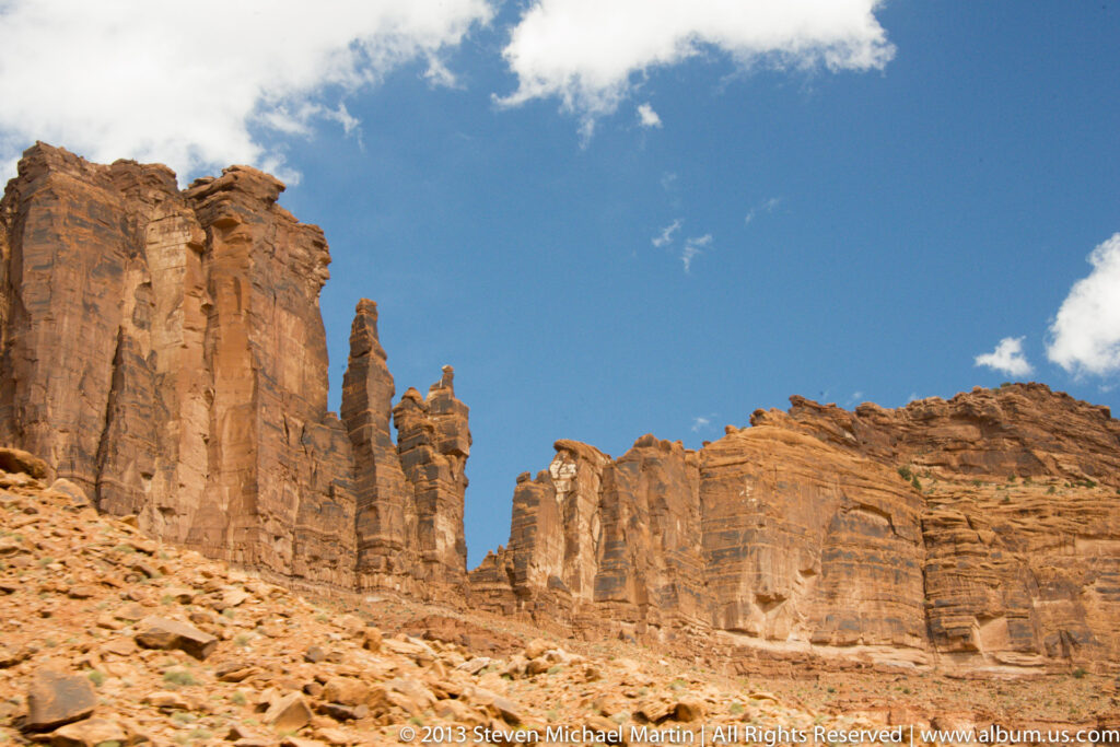 Tall red sandstone towers rise above a broken-rock slope, their narrow pinnacles outlined against a blue sky.