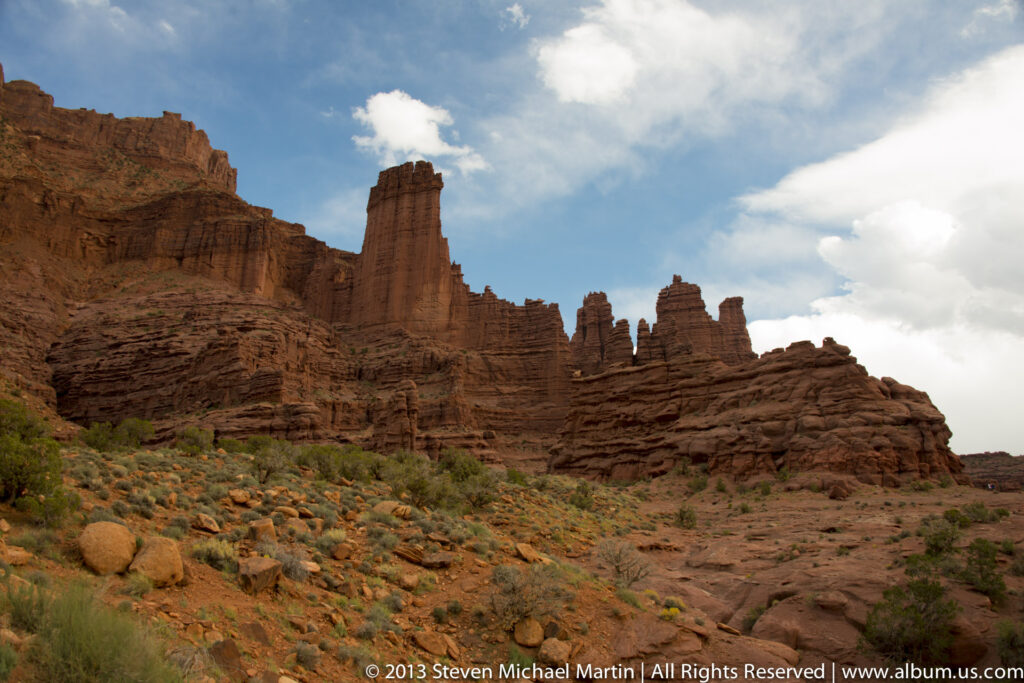 A narrow sandstone tower rises above clustered red spires, with low scrub and loose stones under a cloudy sky.