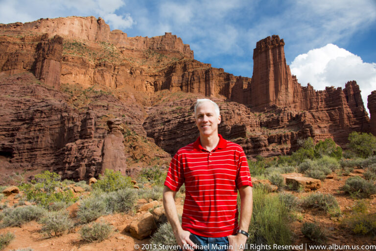 A man in a red striped polo poses before a sandstone tower and layered red cliffs among desert shrubs.