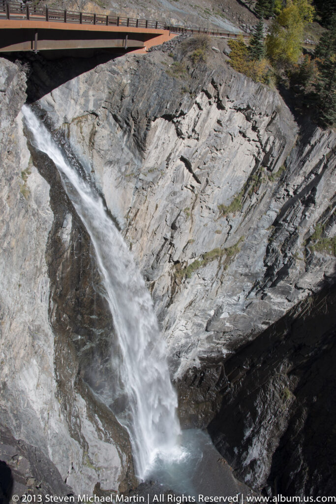 A waterfall plunges through a narrow rock cleft below a pedestrian bridge, flowing over slanted stone into a dark pool.