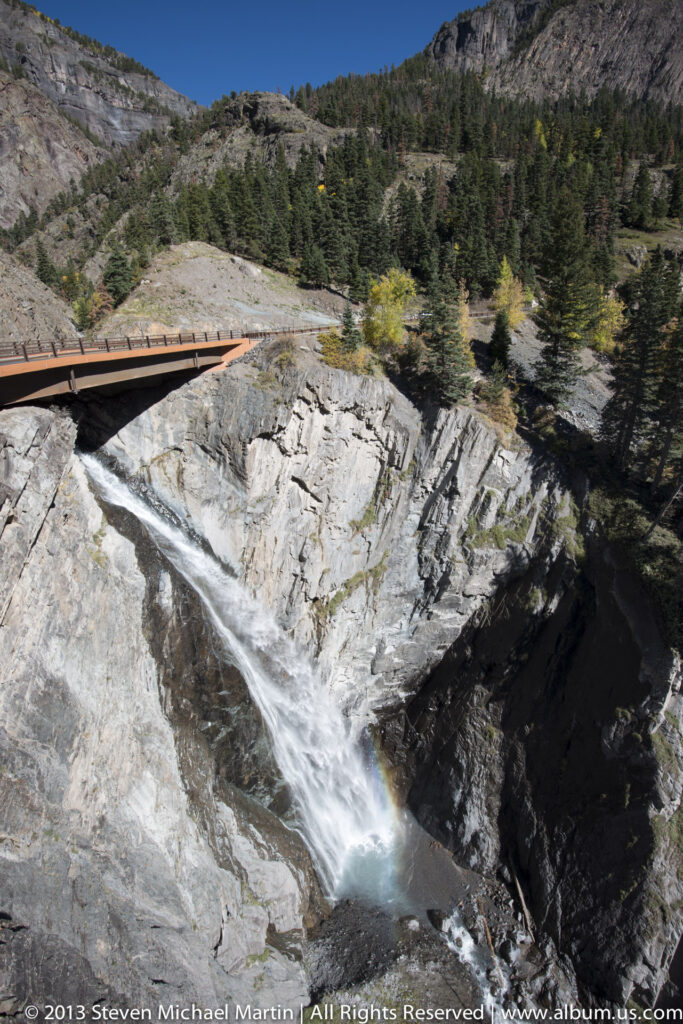 A broad view of a waterfall below a bridge, with pale sloping bedrock, a turquoise pool, and conifers on the rim.
