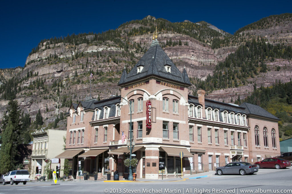 A red-brick hotel with a corner tower and vertical HOTEL sign stands on a street below a steep mountain cliff.