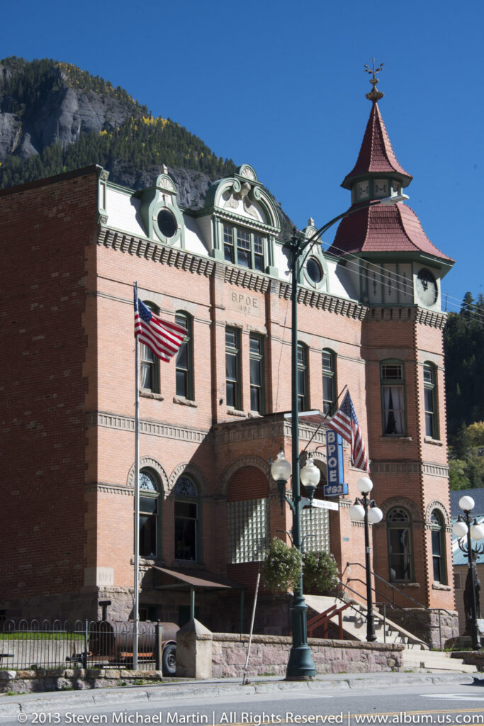 A red-brick façade with a red-roofed tower, arched openings, a blue BRUCE sign, and flags above the sidewalk.