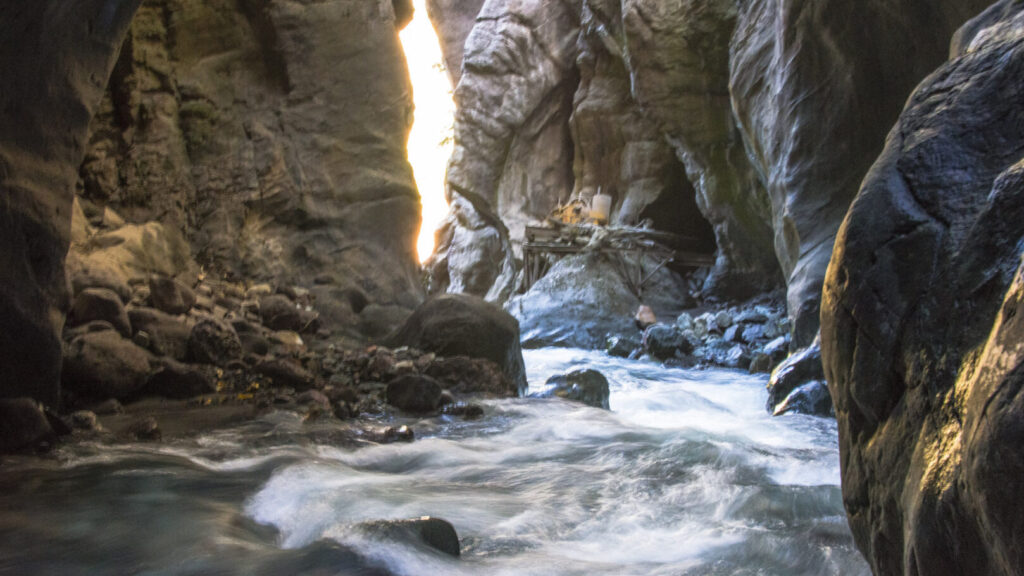Whitewater moves between dark canyon walls beneath a narrow sunlit opening, with a small crossing visible in the distance.