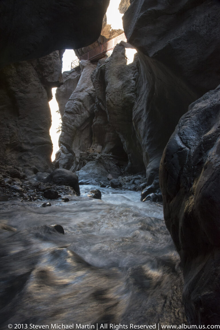 A shaded stream flows around dark boulders beneath an overhang, toward a bright opening crossed by a bridge.