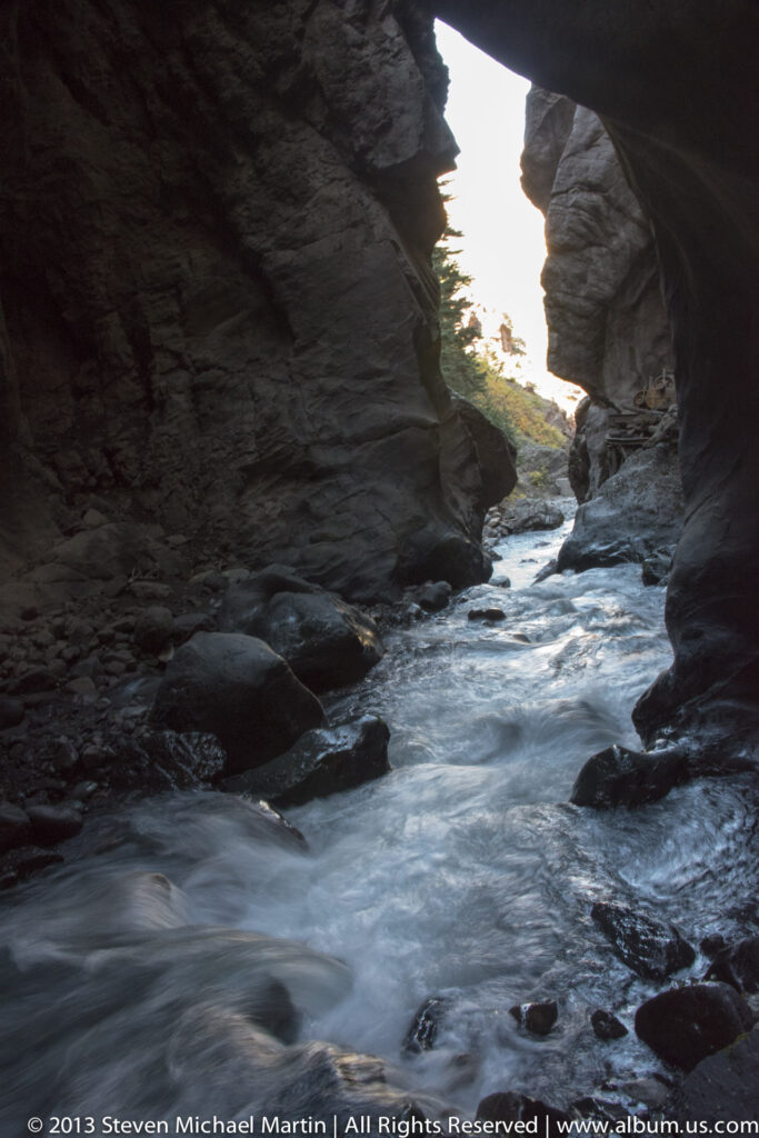 Rippling water runs between shadowed canyon walls toward a narrow opening of bright sky and sunlit trees.