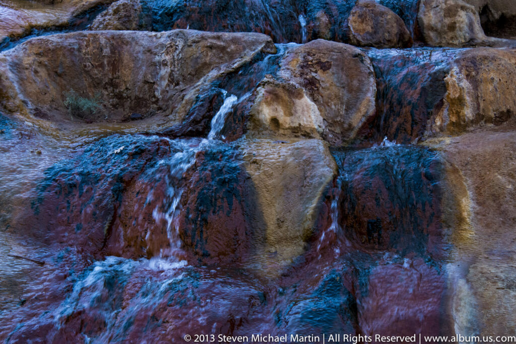 Small cascades flow over rounded rust-colored and gray boulders, with blue reflections across the wet rock.
