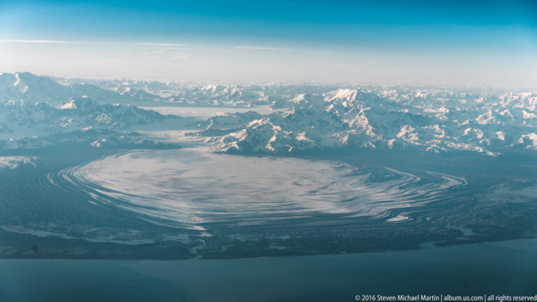 A rounded glacier basin with curved debris bands lies beside blue water beneath a distant snowy mountain range.