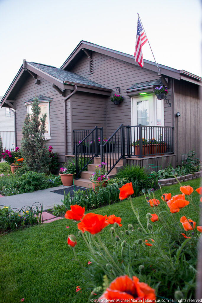 A wood-sided house with an American flag, entry ramp, and flower garden in front.