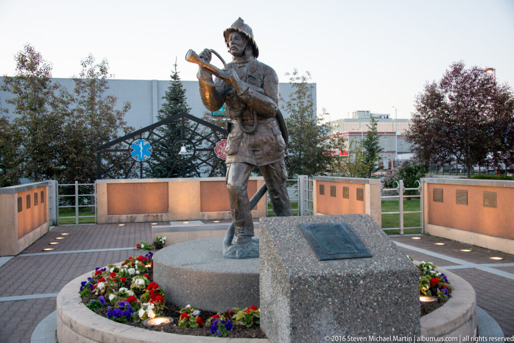 A bronze standing figure faces the camera in a memorial plaza bordered by flower beds and plaque walls.