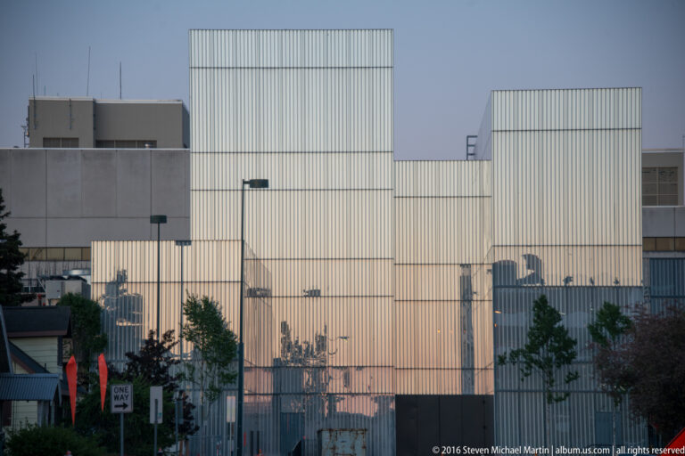 A large corrugated-metal and concrete building stands behind street trees and a one-way sign.