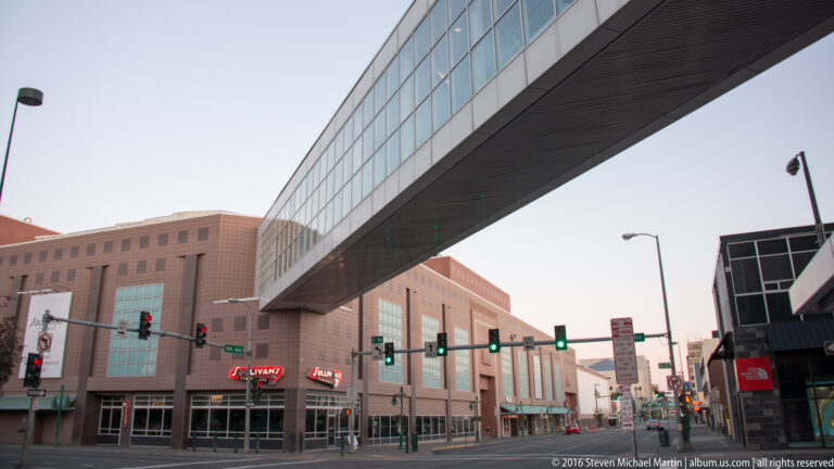 An enclosed skybridge crosses above a downtown Anchorage intersection lined with commercial buildings.