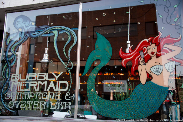 A mermaid illustration and the words Bubbly Mermaid Champagne & Oyster Bar cover a storefront window.