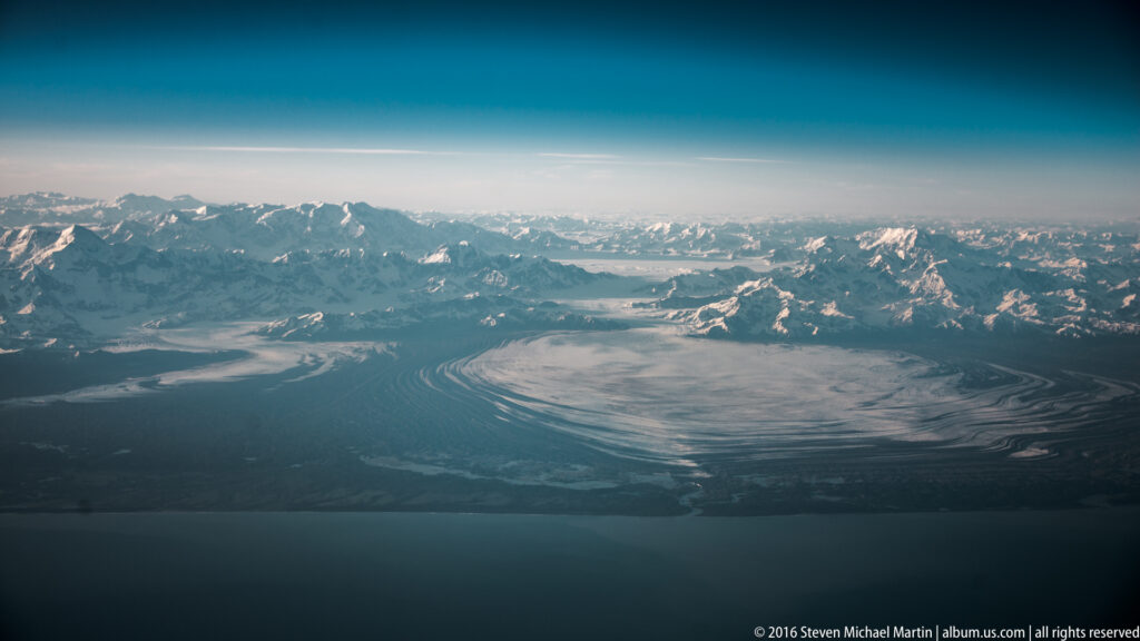 A glacier curves between snow-covered mountain ridges above a blue water body.