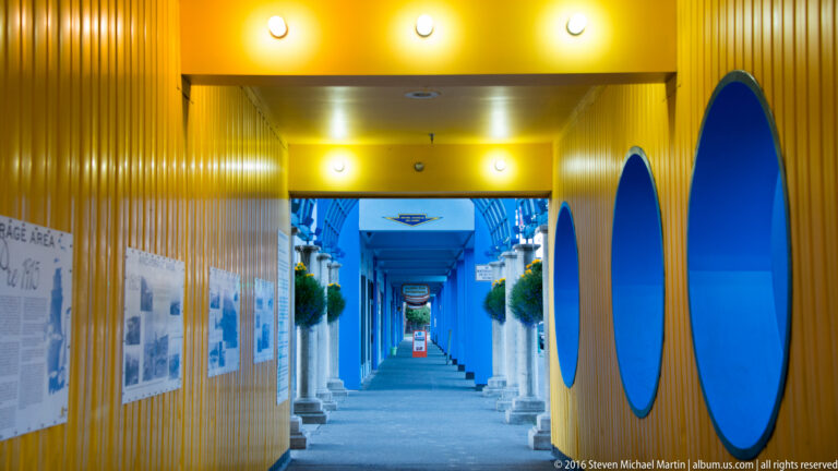 A brightly lit passage has yellow ribbed walls, large blue panels, and a distant blue opening.
