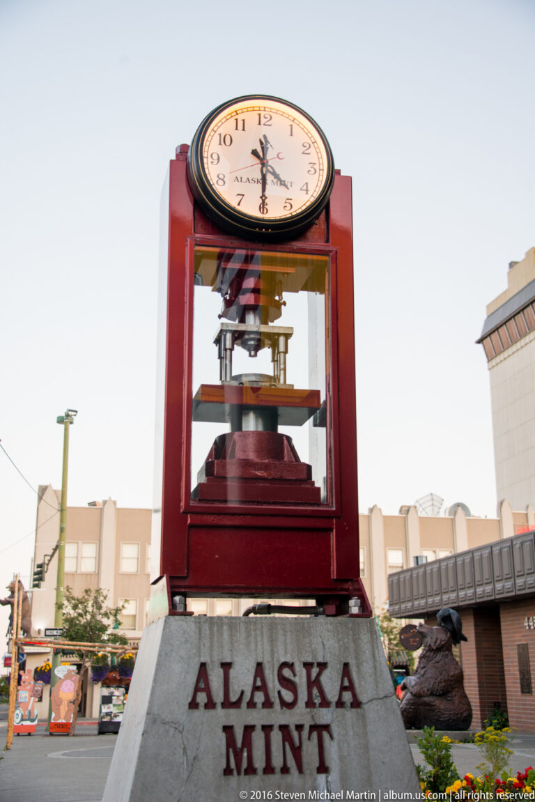 A large clock stands on a red pedestal labeled Alaska Mint beside a downtown street.