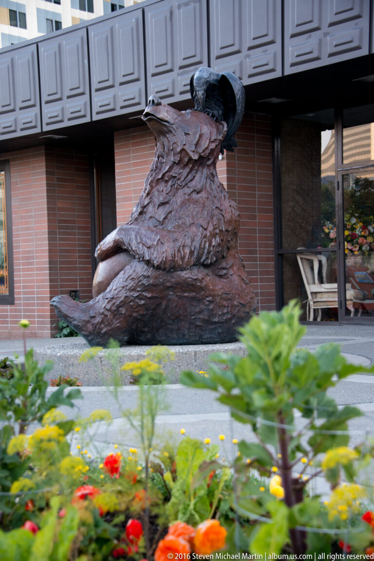 A dark seal-like sculpture rests on a rough stone base in front of a brick building and flower bed.