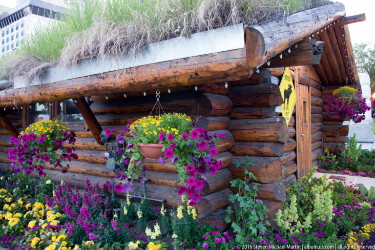 A log building with a grass-covered roof is surrounded by hanging baskets and dense flower beds.