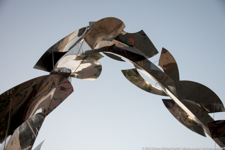 An abstract loop of overlapping reflective metal plates is viewed from below against a clear sky.