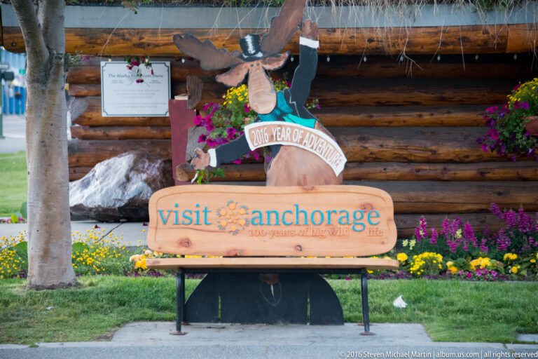 A top-hatted moose character sculpture wears a 2016 Year of Adventure sash beside an Anchorage sign.