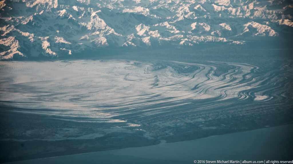 Dark debris bands cross a glacier flowing between snowy mountain slopes toward blue water.