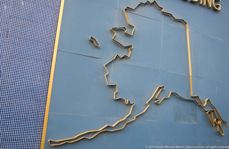 A raised gold outline of Alaska, including the Aleutian Islands, is mounted on a blue-gray wall.