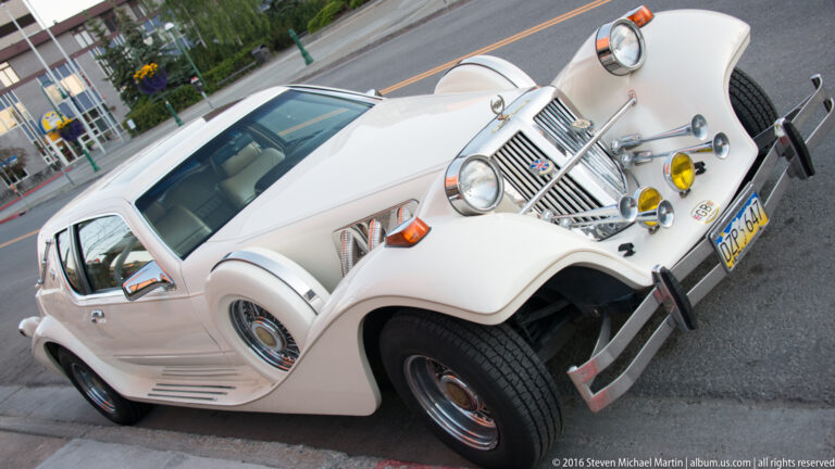 White vintage-style touring car with sweeping fenders, chrome grille, spare tire, and auxiliary lamps parked beside an Anchorage street.