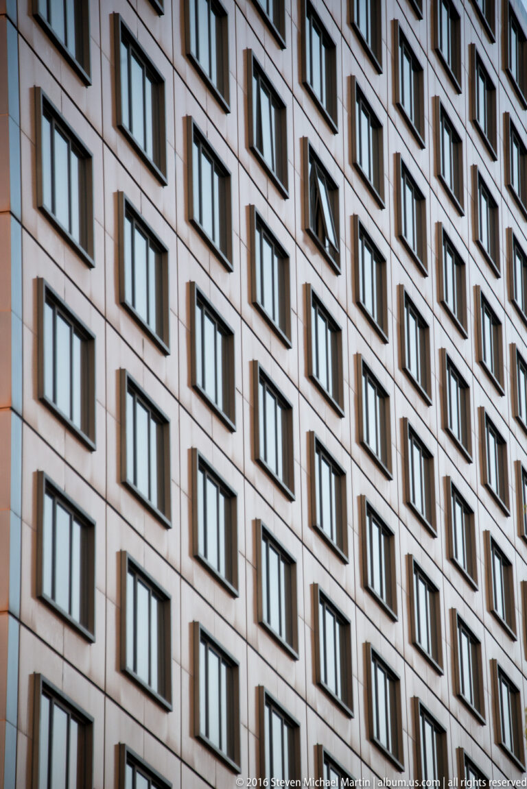 Oblique upward view of a high-rise facade with repeated projecting windows, pale panels, and narrow vertical joints.