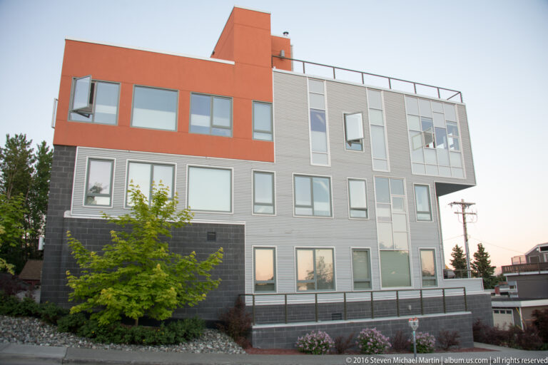 Orange-and-gray multistory building with rows of windows, a rooftop terrace, street trees, and planted frontage.