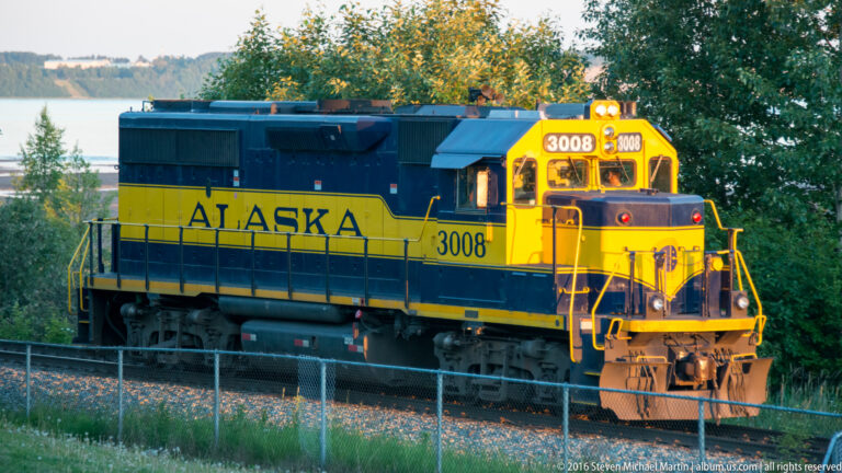 Yellow-and-blue Alaska Railroad locomotive 3008 beside a chain-link fence, with trees and water behind the tracks.