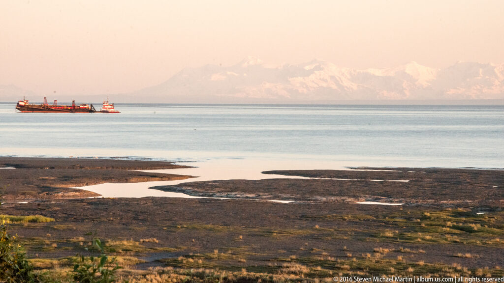 Exposed tidal flats and shallow channels border a coastal inlet, with a vessel and hazy snow-covered mountains beyond.