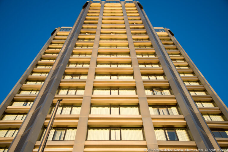 Low-angle view looking up a modern high-rise with repeating window bands and vertical ribs against blue sky.