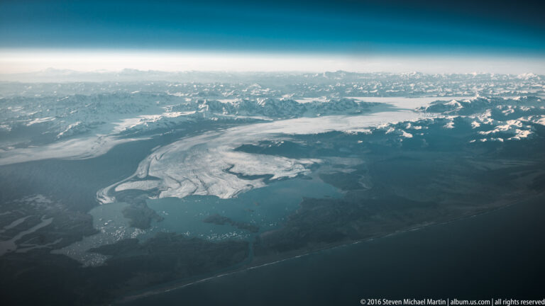 A broad glacier flows from snowy mountains into braided meltwater channels and a dark, forested plain.