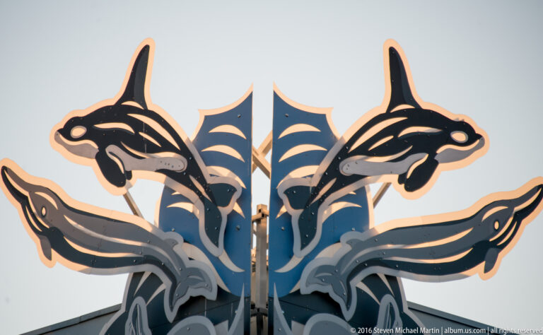 Paired stylized orca figures meet at a central panel in a dark and pale metal wall ornament.