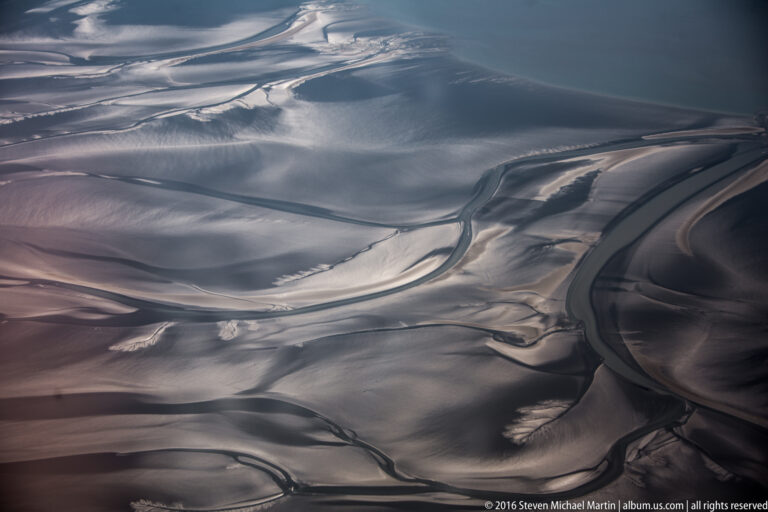 Aerial view of pale braided channels winding across a dark sediment plain with gravel bars and silt patterns.