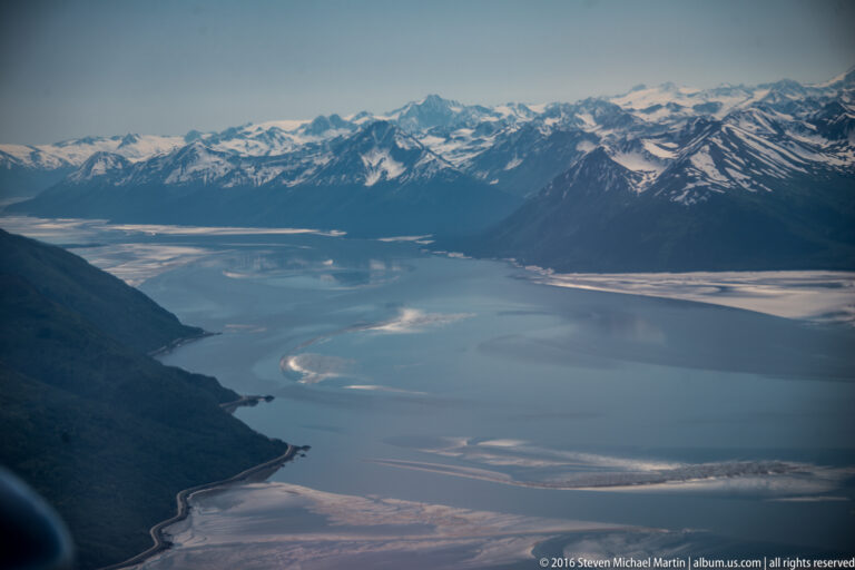 Aerial view across a blue inlet toward snow-streaked mountains, with forested slopes and pale coastal channels.