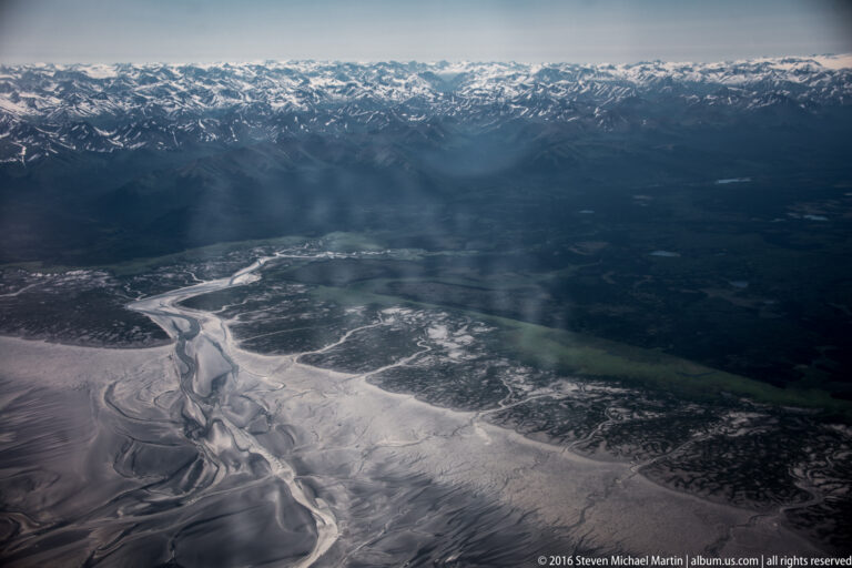 Turquoise meltwater pond set in a snowfield between dark rocky ridges, viewed from above.