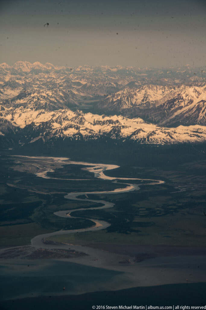 Braided silver channels wind across a shadowed plain below a long, snow-covered mountain range.