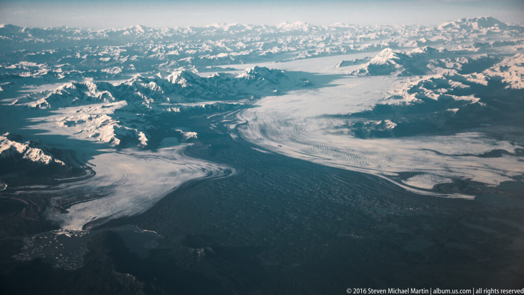 Several glacier branches converge through a snowy mountain valley above a dark lowland and water edge.
