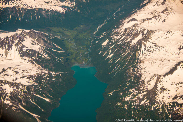 Long turquoise lake lies between steep snow-covered mountains, narrowing toward a green valley at its head.