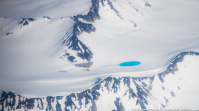 Bright blue meltwater pool set in pale snow and ice beside dark rocky outcrops.