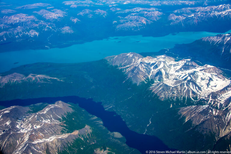 Aerial panorama of blue lakes among snow-streaked ridges and dark forested mountain slopes.
