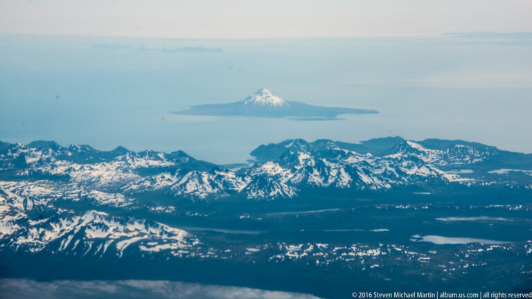 Snow-capped conical peak rises beyond rugged nearer mountains, with blue coastal water and haze between them.
