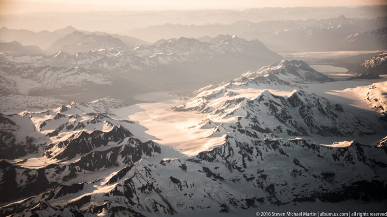 Golden light falls across snowy mountain peaks and glaciers seen from above at a low angle.